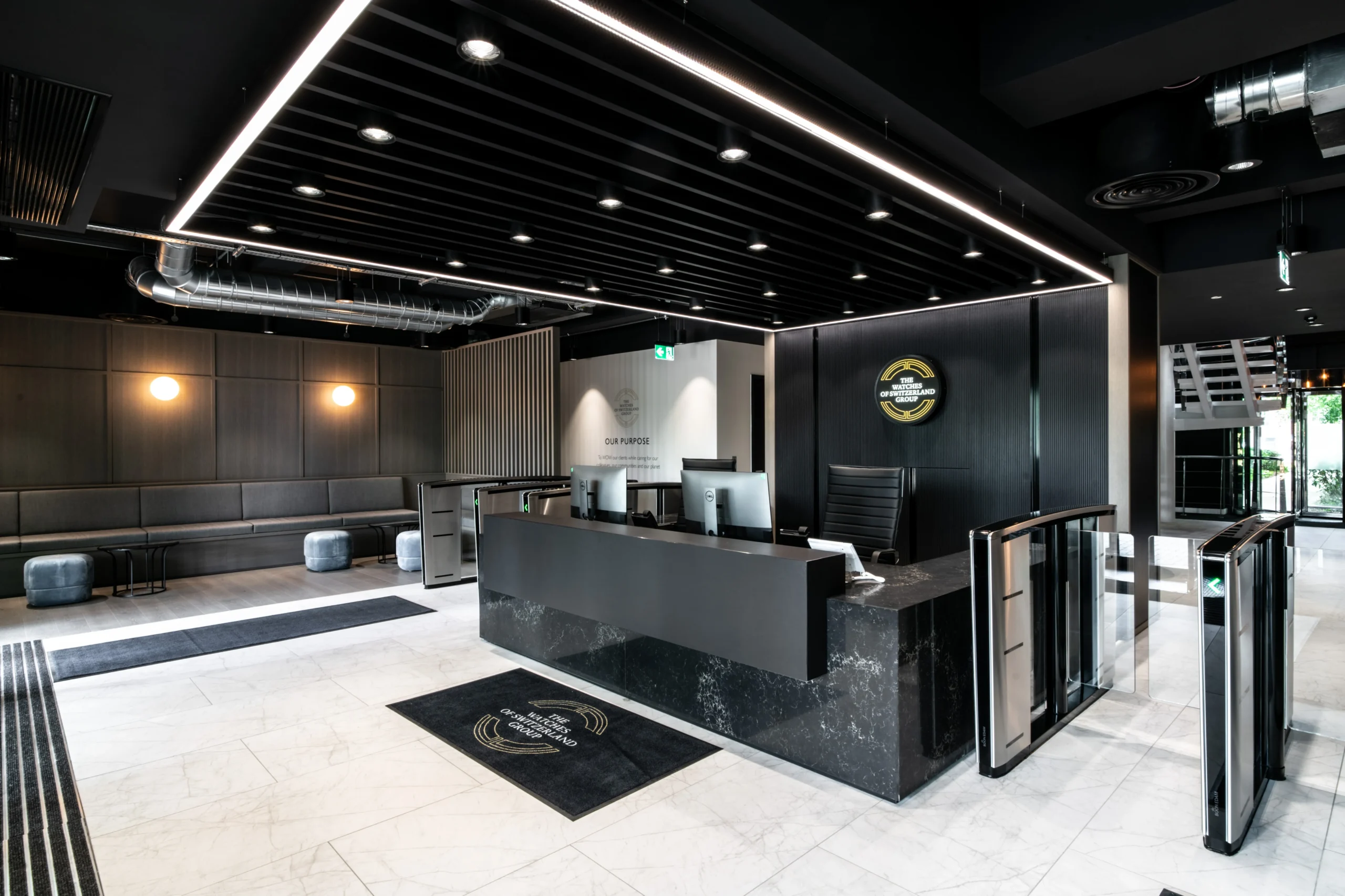 Reception at Watches of Switzerland with black marble desk, branded signage, soft bench seating and crisp monochrome finishes throughout
