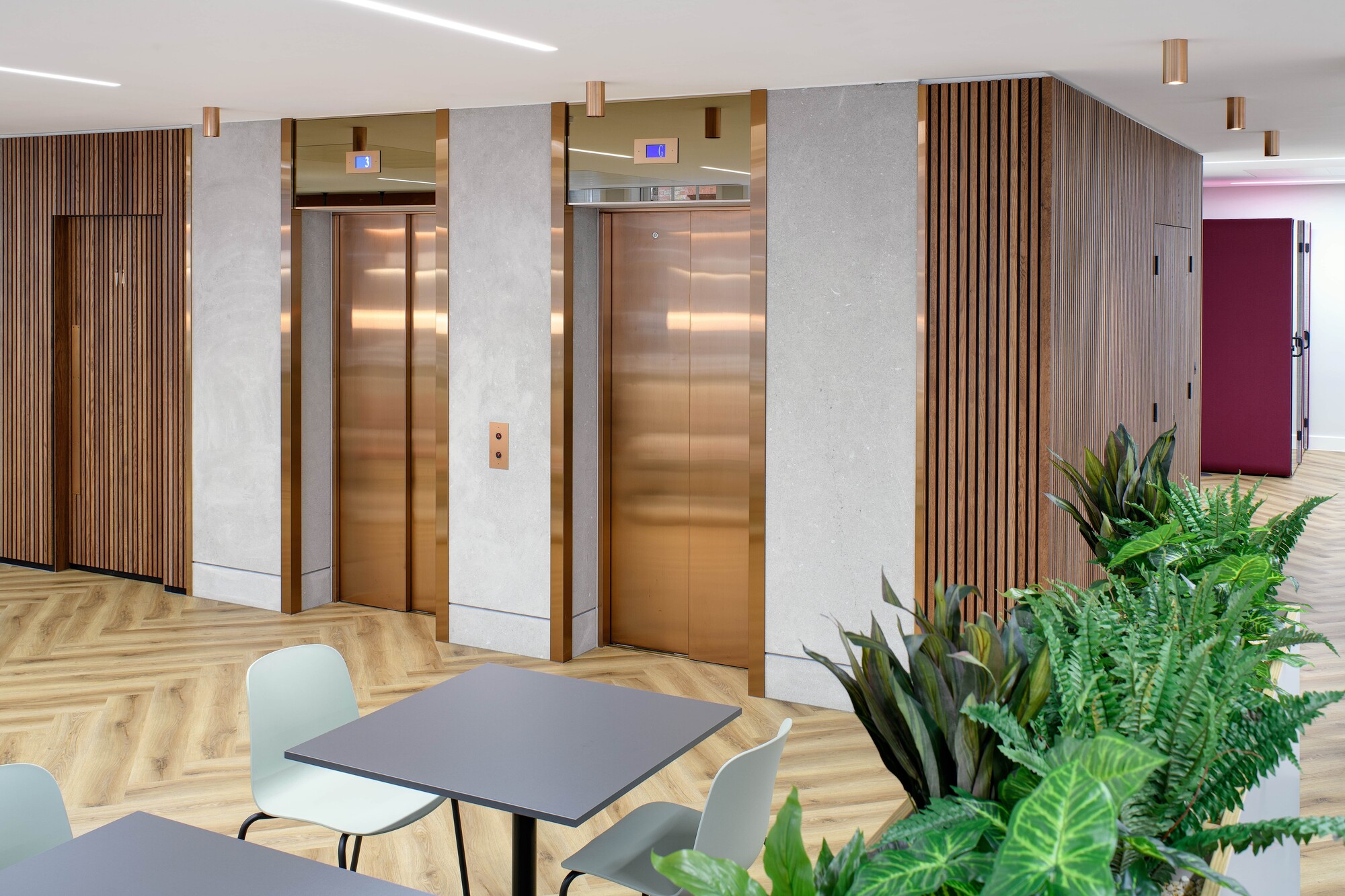 Tenant-Ready™ breakout space for an Asset Management Firm, with café tables and chairs placed near gold-toned lift surrounds, wood panelling, and indoor planting.