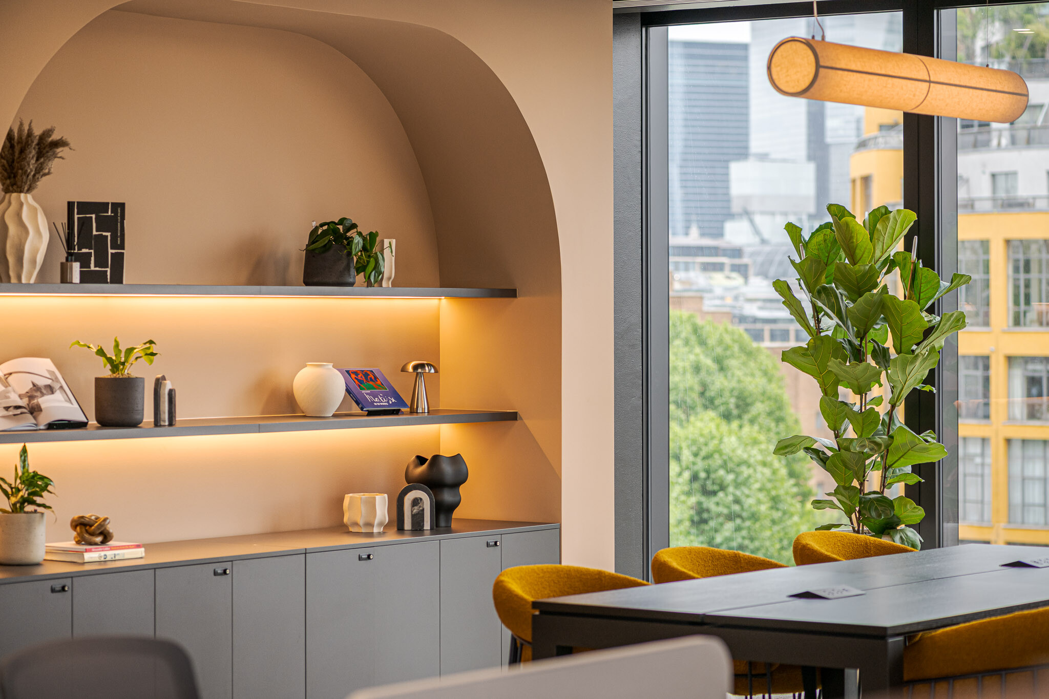 Detail view of Arbor’s Tenant-Ready™ breakout area, furnished by Platfform, featuring backlit shelving, sculptural décor, mustard chairs and a large window with views over London.