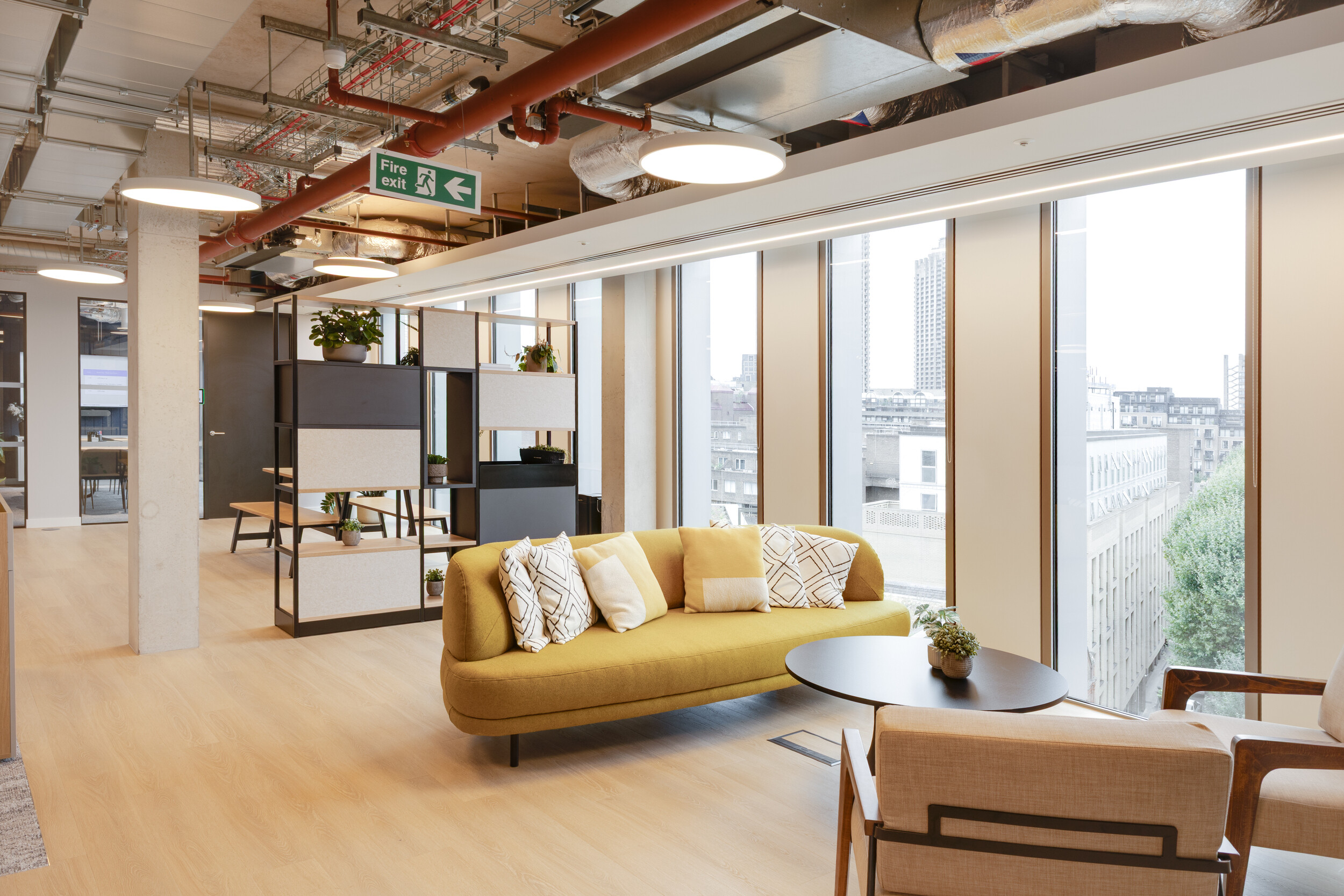 Capital on Tap lounge with a mustard yellow sofa and patterned cushions, paired with neutral armchairs and a black round table, framed by shelving with plants.