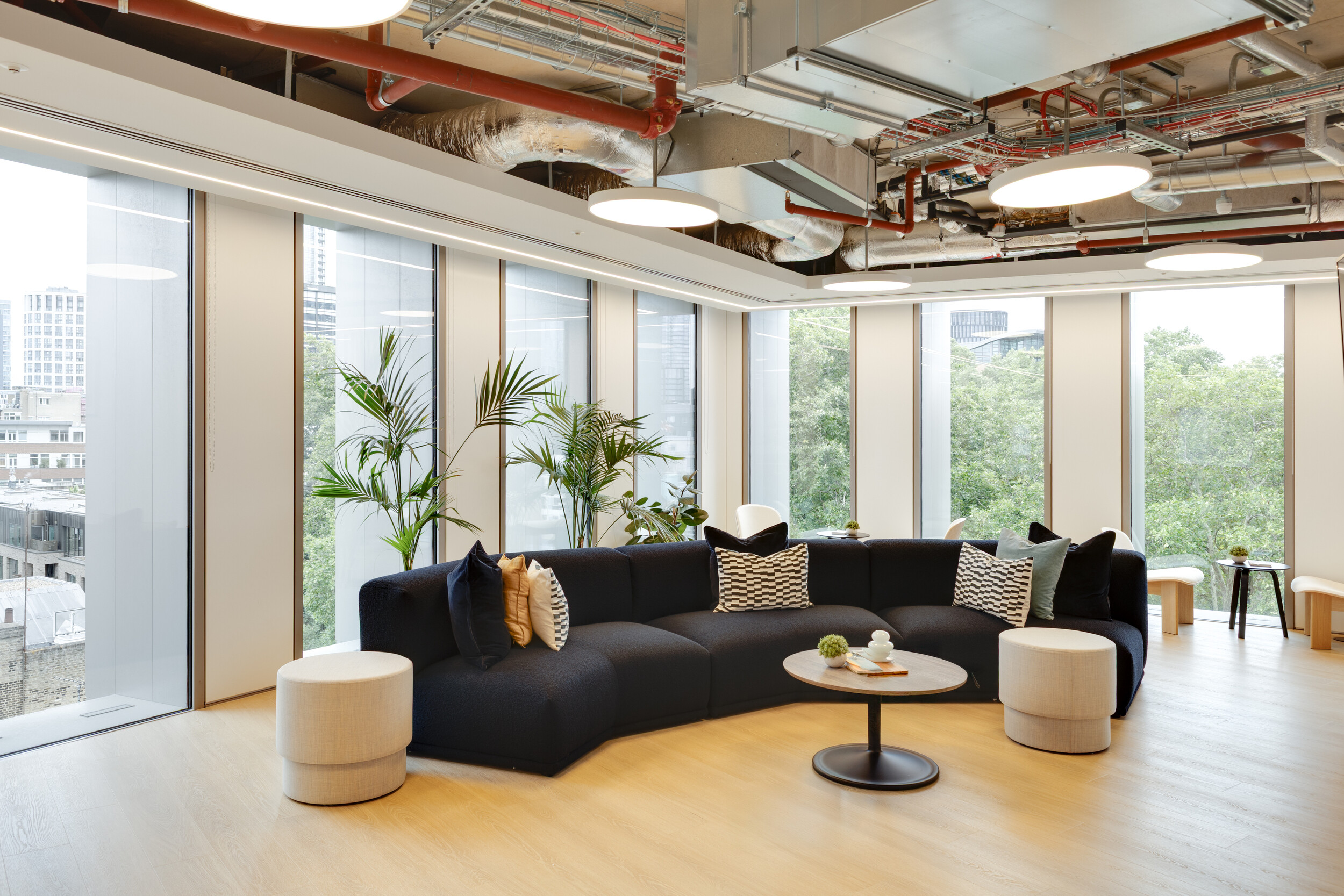 Capital on Tap lounge with a curved navy sectional sofa, patterned monochrome cushions, and round neutral stools, complemented by a black coffee table and tall potted plants.