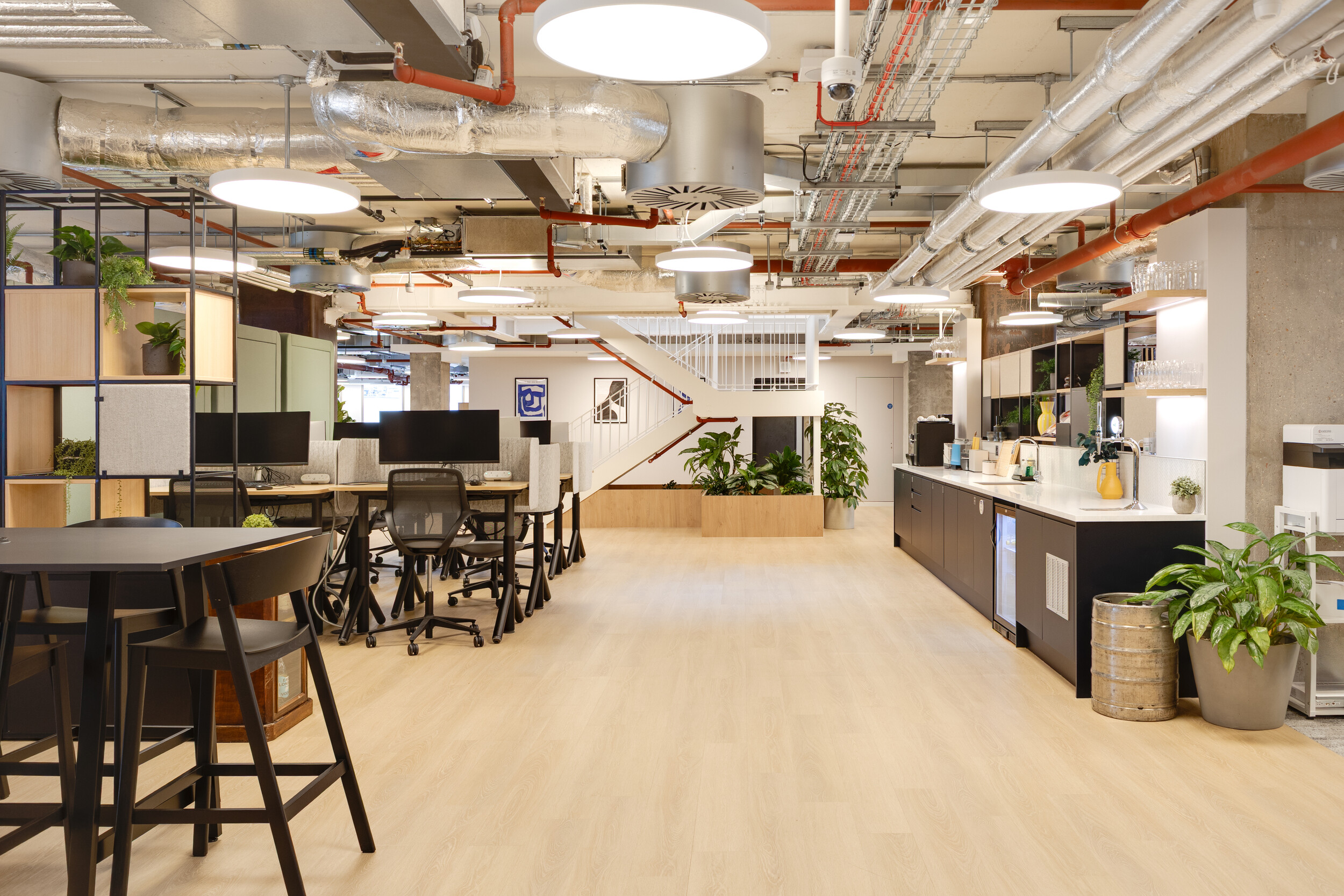 Capital on Tap workspace with a teapoint featuring dark cabinetry and open shelving, high tables with black stools, and shared desks near a central staircase and greenery.