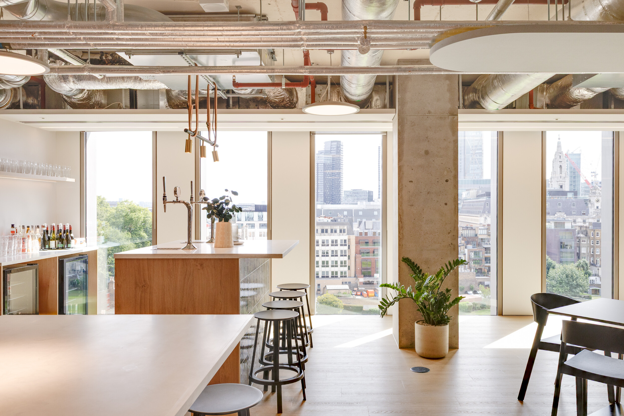 Capital on Tap bar area with tall black stools, wood counters, and glass-front fridges, styled with greenery and warm neutral tones against floor-to-ceiling windows.