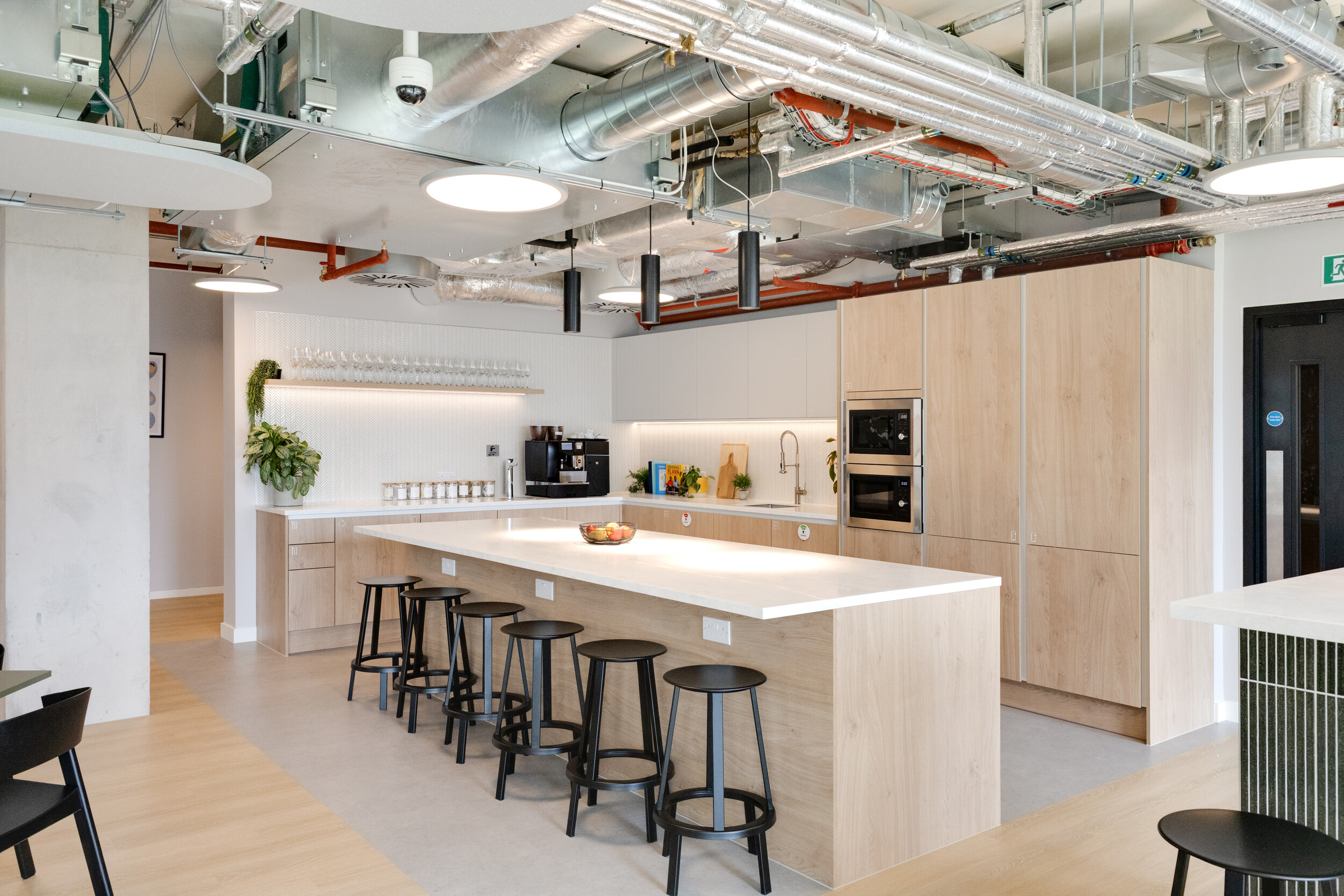 Capital on Tap kitchen with a long light wood island, black bar stools, and integrated cabinetry in pale tones, styled with greenery and open shelving for a modern finish.