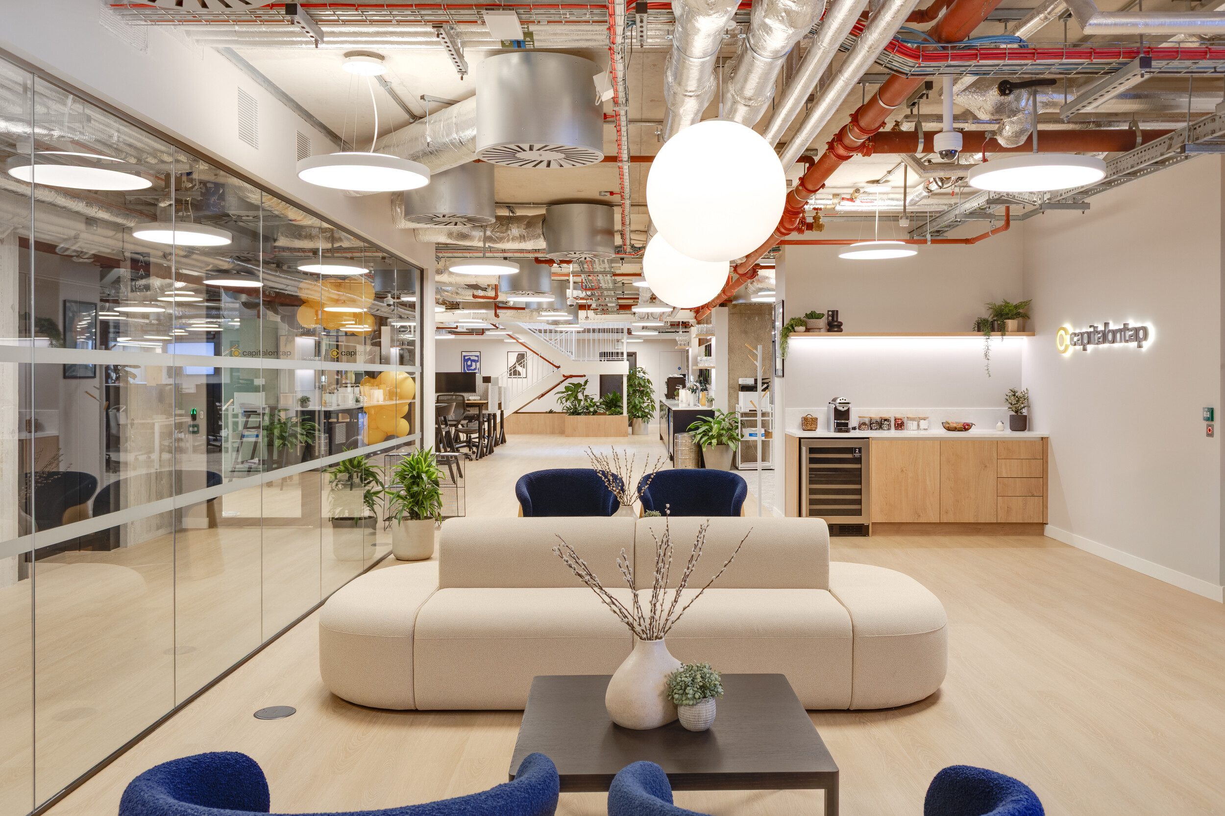 Capital on Tap lounge with a cream curved sofa, deep blue armchairs, and light wood cabinetry, styled with plants and modern lighting for a welcoming workspace.
