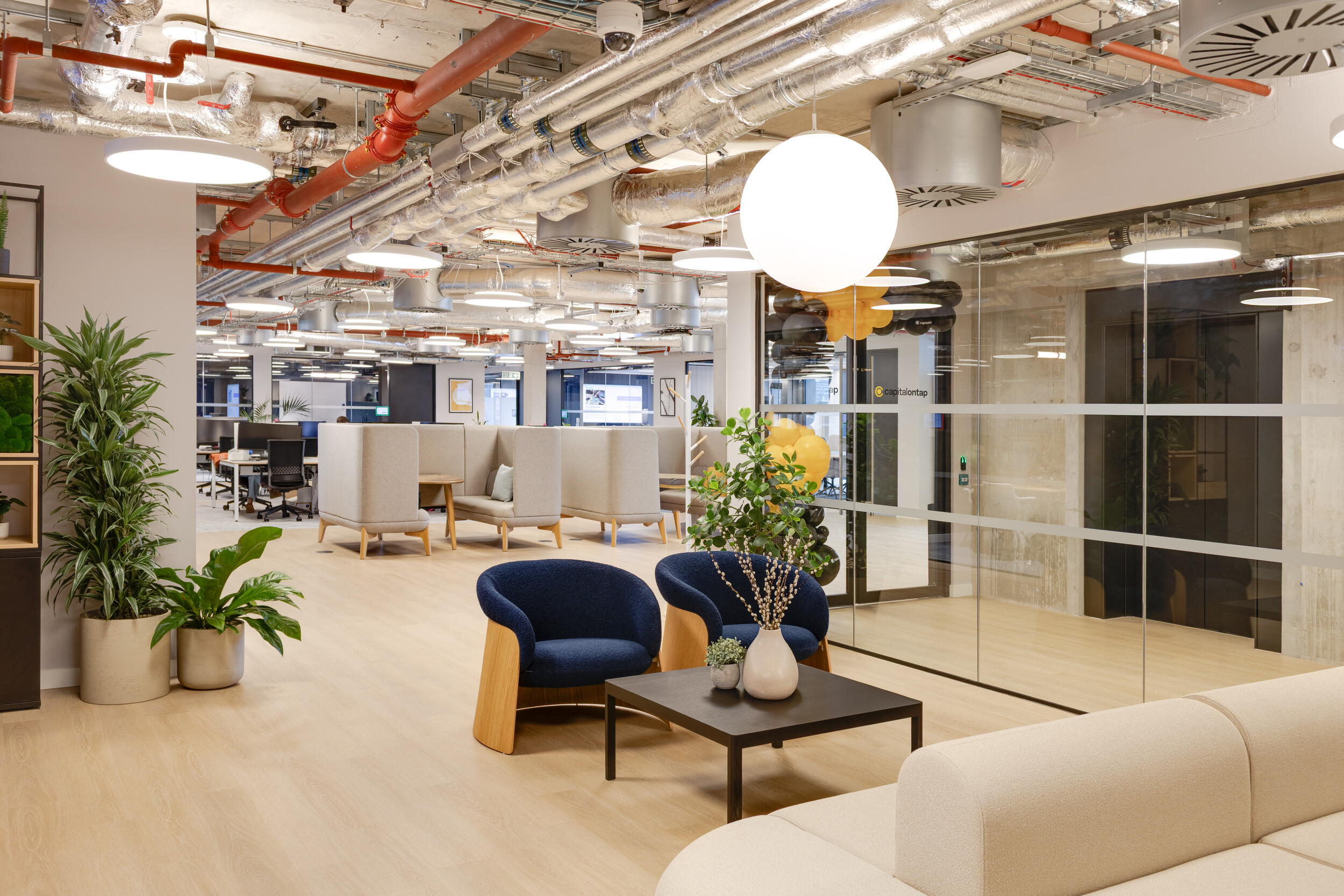 Capital on Tap office lounge with navy armchairs and a black coffee table, set beside cream modular seating and private booth-style work areas in soft grey fabric.