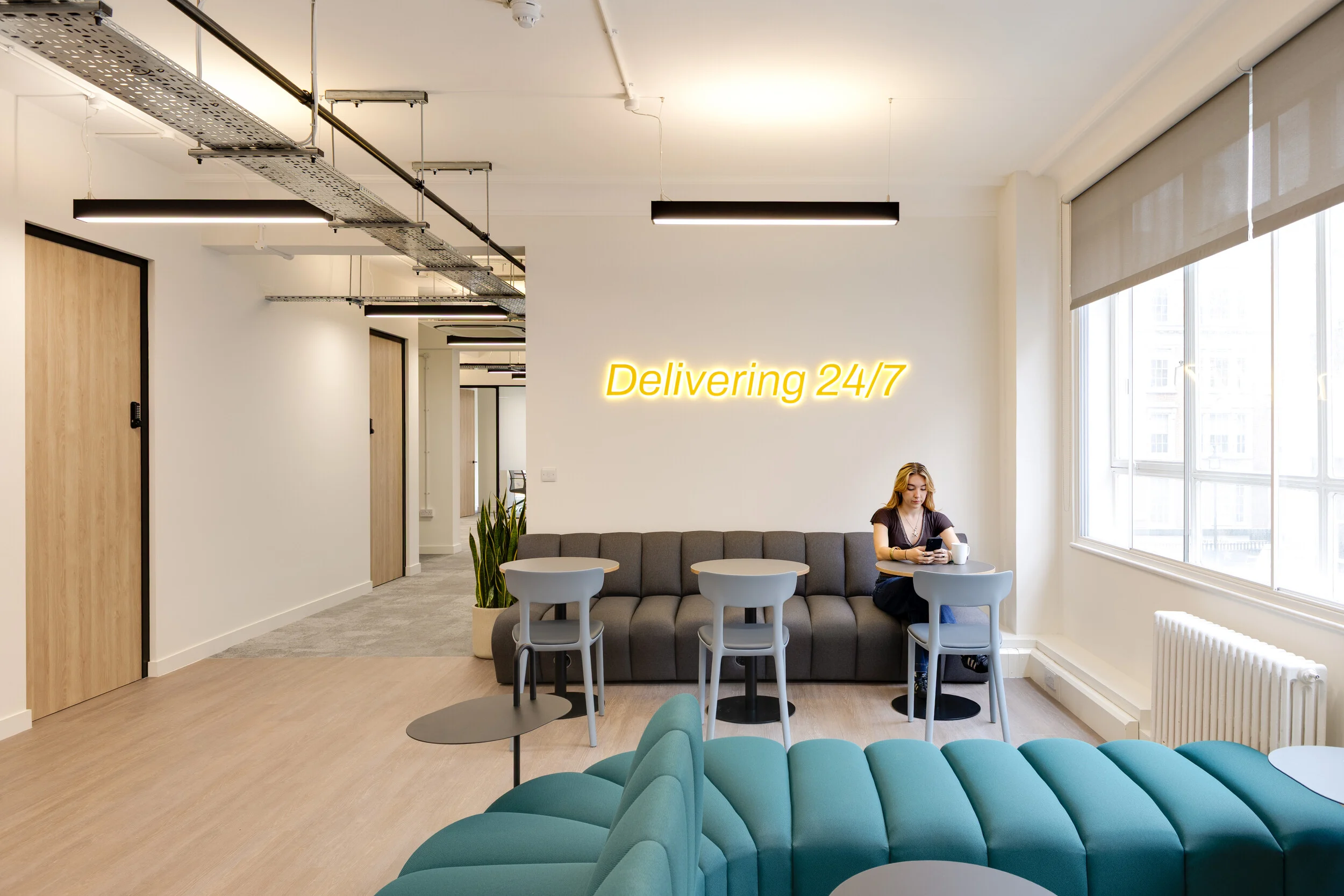 A breakout area at InPost’s London office furnished by Platfform, with grey seating, café tables, and a neon “Delivering 24/7” sign. The space is bright and relaxed, designed for casual moments between work.